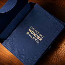 A dark blue textured book or journal with gold foil text reading 'LIFE WITHOUT WONDERS IS NO LIFE AT ALL' lies open on a warm wooden surface.