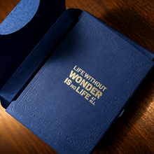 A dark blue textured book-style box with a matching inner sleeve, open on a wooden surface. The cover has gold foil text reading 'LIFE WITHOUT WONDER IS NO LIFE AT ALL'.