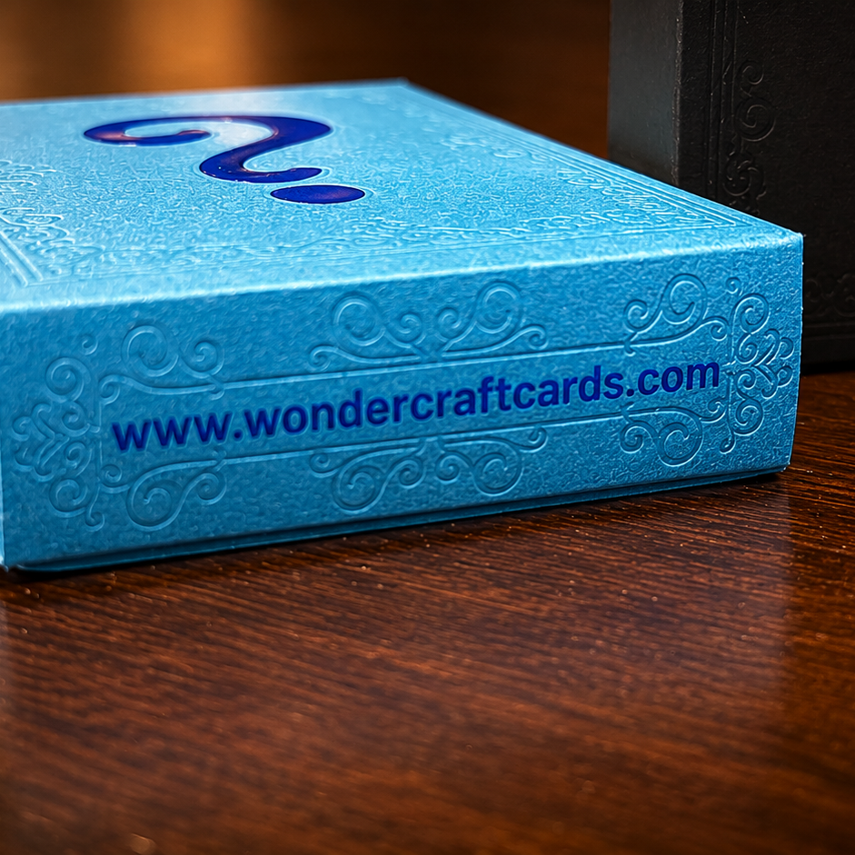 A bright blue rectangular box with embossed decorative patterns and a dark blue question mark logo on top, sitting on a wooden surface. The front of the box shows the website 'www.
